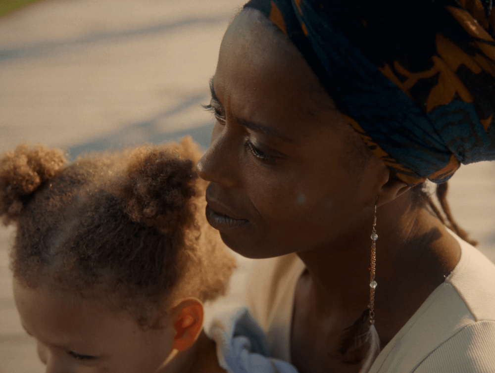 A woman with a headwrap looks thoughtfully while holding a young child.